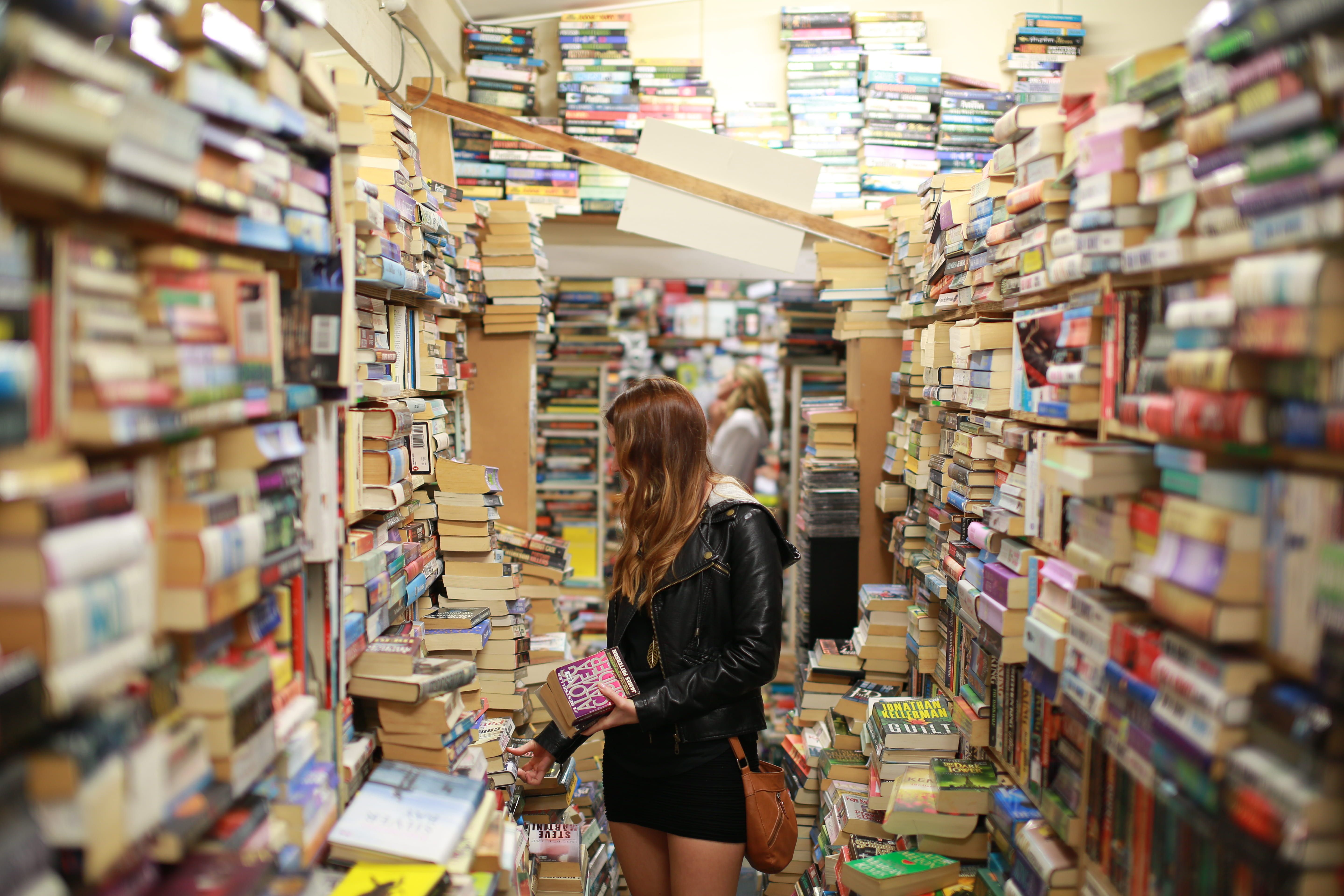 A woman stands between bookshelves with piles of books at a used bookstore.