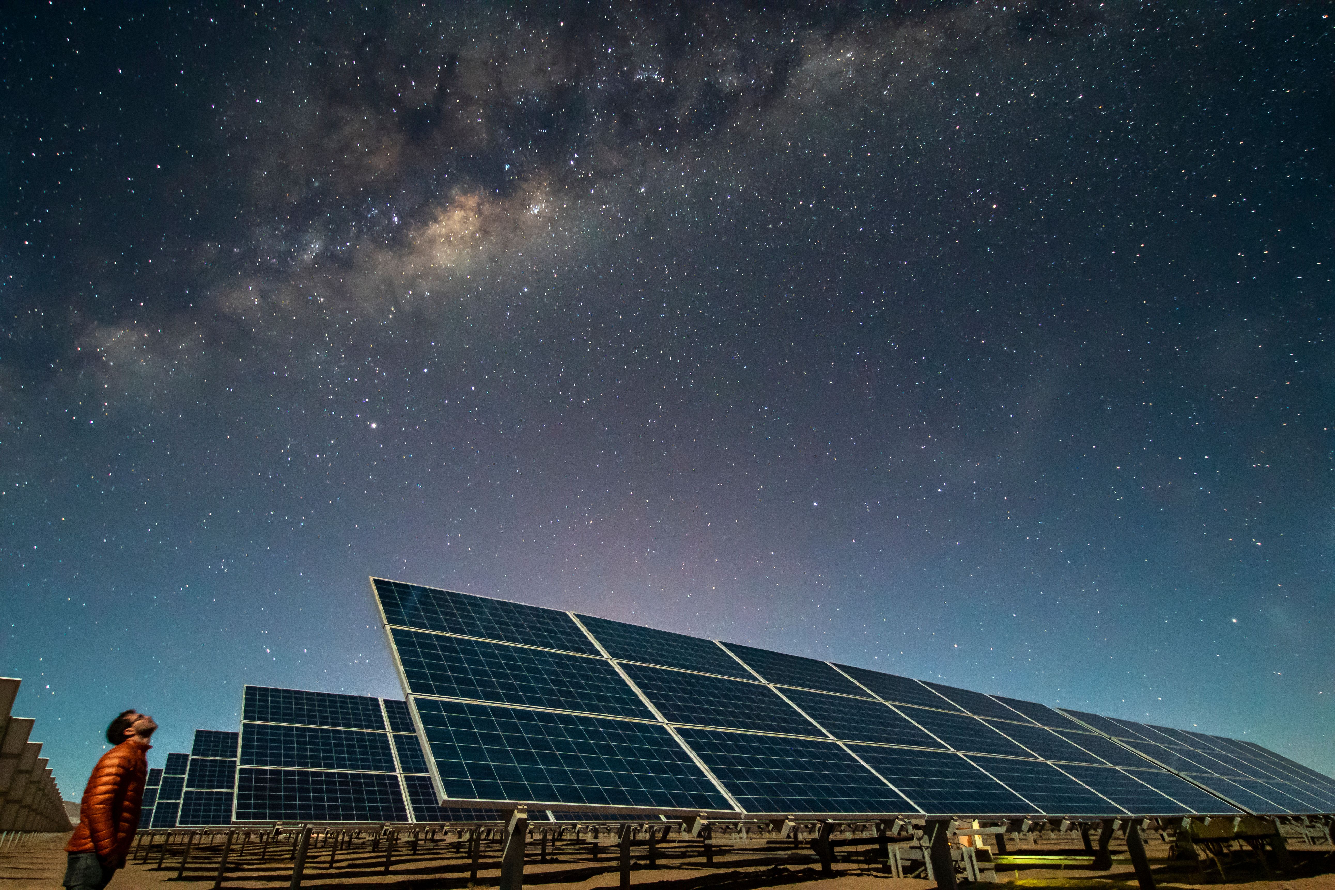 A man stands next to large groundmount solar panels and looks up at the night sky.
