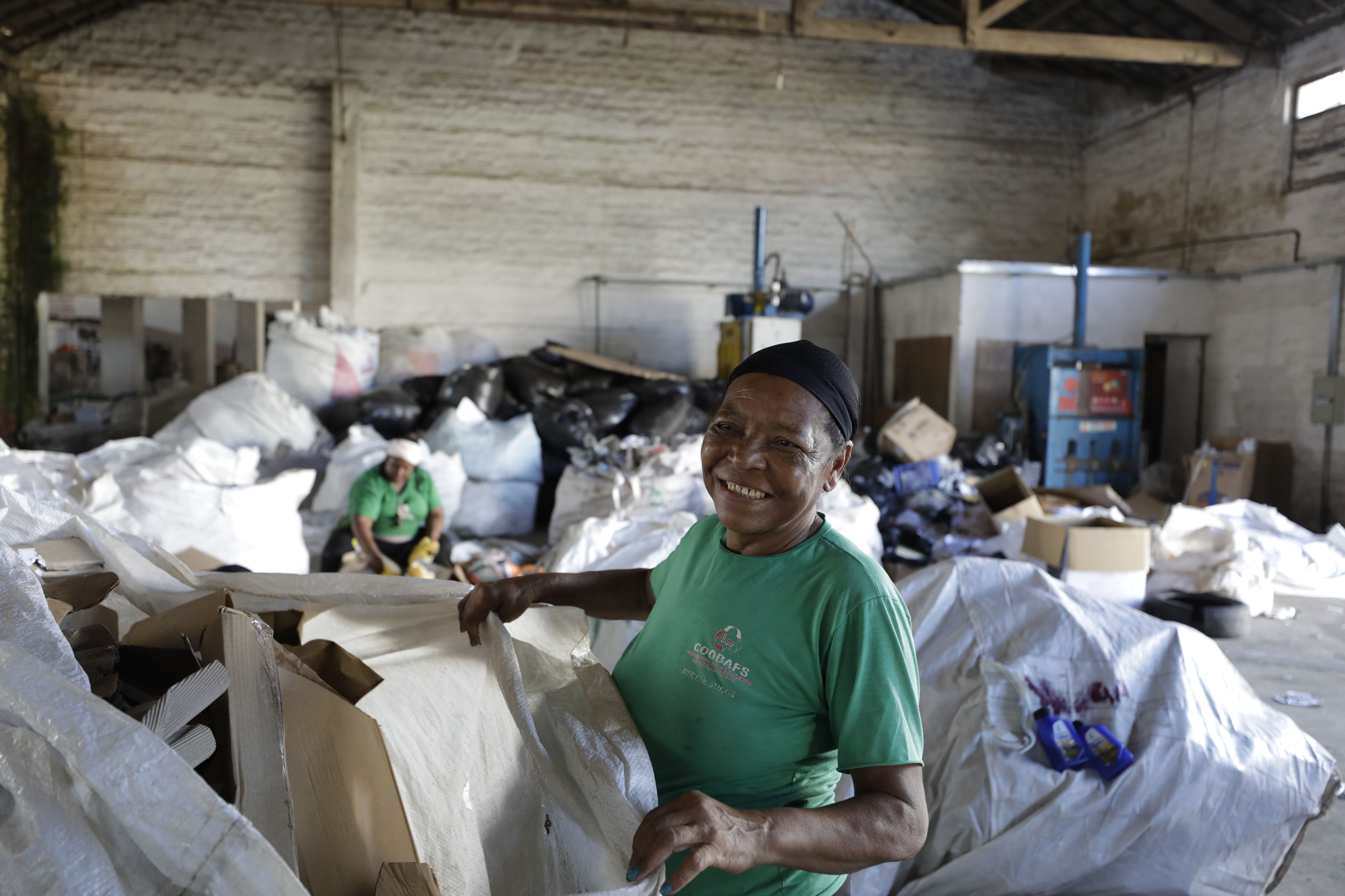 A worker at a recycling cooperative in Brazil smiles as she sorts waste. Another workers is sorting waste in the background and there are several large white bags stuffed with waste behind her. 