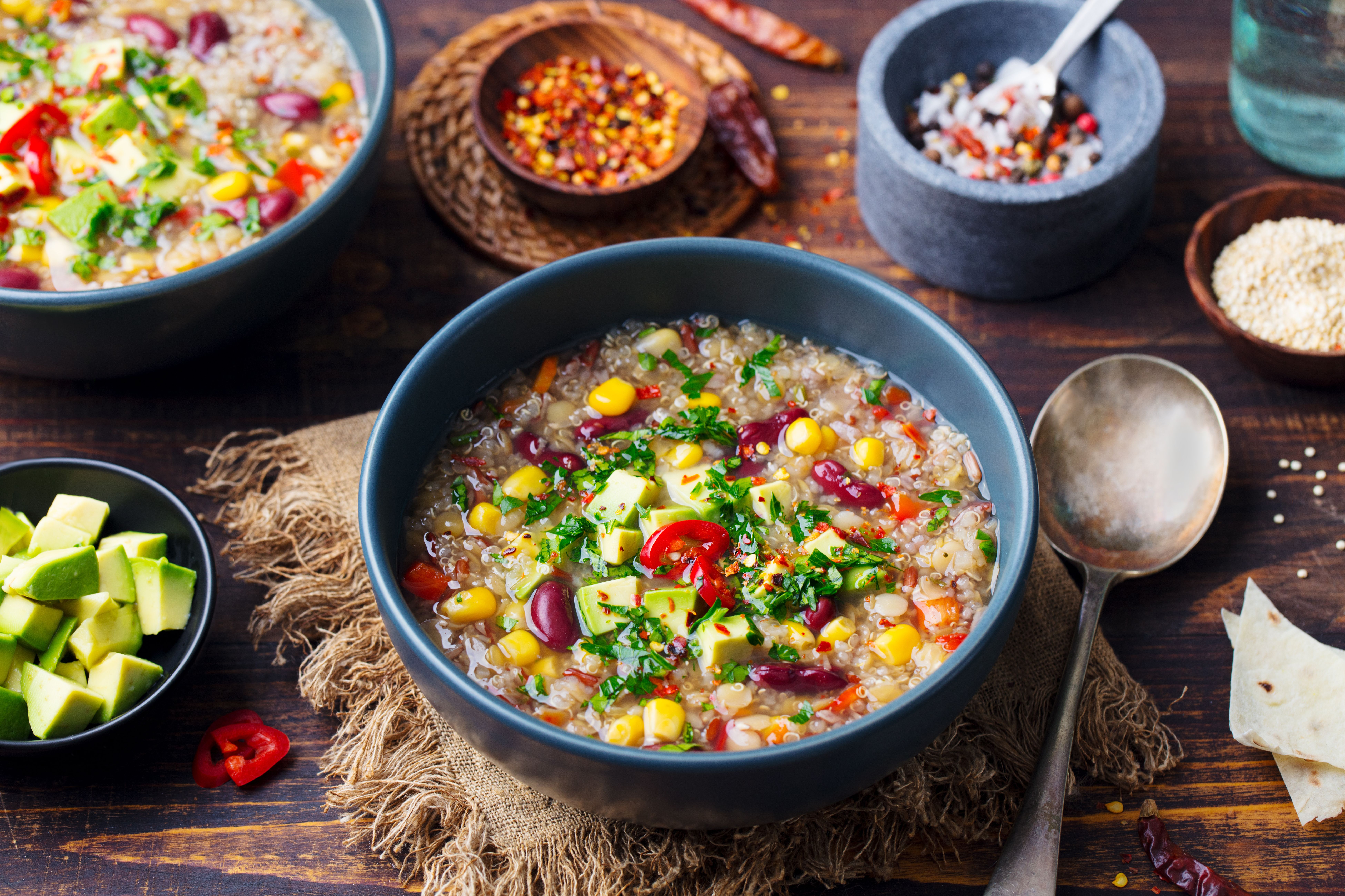 A bowl of colorful soup with quinoa, red beans, corn, avocado, and green garnish.